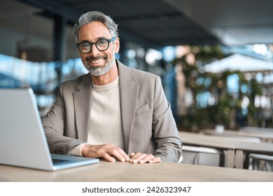 Happy confident mature professional business man hybrid working looking at camera. Smiling middle aged businessman wearing glasses sitting at outdoor office table using laptop. Portrait. Copy space. - Powered by Shutterstock