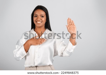 Happy confident millennial black businesswoman in white blouse presses hand to chest and swears, isolated on gray background, studio. Gratitude, promise, take an oath in business, work and startup