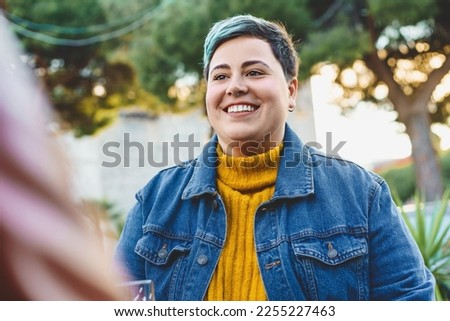 A happy and confident non-binary curvy woman sits in an outdoor bar. Smiling in this half-body portrait embracing her unique identity and celebrating it. self-acceptance, body positivity and diversity