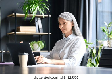 Happy Confident Older Senior Asian Businesswoman Ceo Executive Manager Sitting At Desk Working Typing On Pc Laptop Computer In Contemporary Corporation Office. Business Technologies Career Concept.