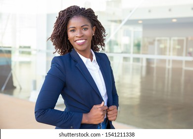 Happy confident professional posing near office building. Young African American business woman standing outside, adjusting formal jacket, looking at camera, smiling. Successful businesswoman concept