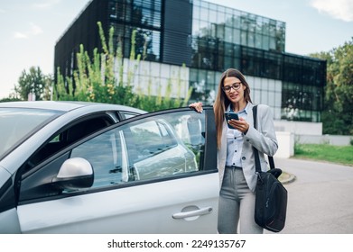 Happy confident professional woman entering a car on a driver seat near office building. Young business female using smartphone and wearing a suit and a business bag. Successful businesswoman concept. - Shutterstock ID 2249135767