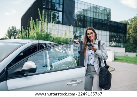 Happy confident professional woman entering a car on a driver seat near office building. Young business female using smartphone and wearing a suit and a business bag. Successful businesswoman concept.