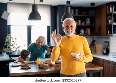 Happy confident senior man looking at camera and smiling showing with hand ok sign satisfied with breakfast porridge, standing in front of family members, senior woman and two kids in the kitchen. - Powered by Shutterstock