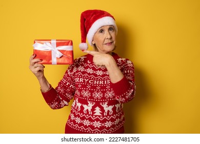 Happy confident senior woman wearing red santa hat stands with gift box and looking at the camera isolated on yellow. Mature lady promoting new year deal, holiday sale - Powered by Shutterstock