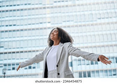 Happy confident successful professional young African American business woman office leader executive wearing suit outstretching arms standing in big city street feeling success and freedom outdoors.