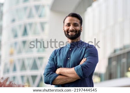 Happy confident wealthy young indian business man leader, successful eastern professional businessman crossing arms looking at camera posing outdoors in urban big city for close up headshot portrait.