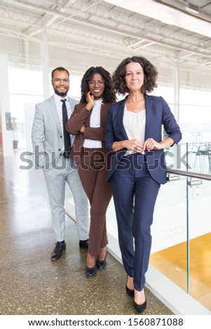 Happy confident workgroup posing for camera in contemporary office hallway. Business man and women standing together, looking at camera, smiling. Successful creative group concept