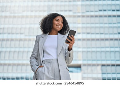 Happy confident young African American business woman using cell mobile phone outdoor. Smiling lady professional holding cellphone standing at city street building looking away working outside office.