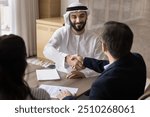 Happy confident young Arabian company owner shaking hands with European colleagues, buyers, clients, talking to business partners at meeting table, giving handshake, smiling, laughing