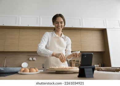 Happy confident young baker woman shooting preparing pizza on cellphone in home kitchen, dropping cheese on dough at smartphone fixed in holder, smiling, laughing - Powered by Shutterstock