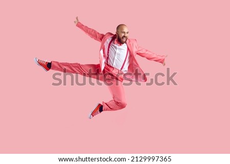 Happy confident young bald man jumping in the studio. Full body shot of a funny joyful male dancer wearing a party suit, bowtie and trainer shoes jumping high in the air isolated on a pink background