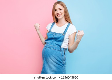 A Happy Confident Young Beautiful Pregnant Woman In A Denim Jumpsuit Shows A Winning Gesture While Standing On An Isolated Pink And Blue Background