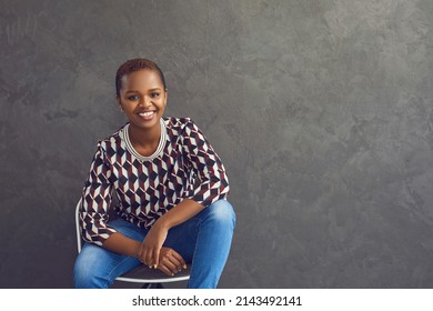 Happy confident young black woman sitting in relaxed pose on chair against grey copyspace background. Portrait of successful African American fashion brand owner. Female success and leadership concept