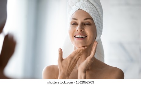 Happy Confident Young Lady With Towel On Head Look In Bathroom Mirror Touching Moisturized Soft Healthy Sensitive Skin Doing Morning Hydration Spa Beauty Routine, Facial Skincare Treatment Concept