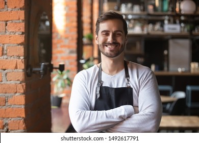 Happy confident young waiter entrepreneur looking at camera, smiling male small cafe business owner employee standing in restaurant, millennial businessman wear apron posing in coffee shop portrait