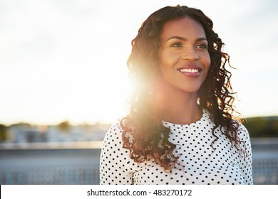 Happy confident young woman backlit by the morning sun standing on an urban rooftop looking into the distance with a lovely warm smile