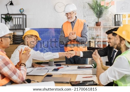 Happy construction team shaking hands during conference at office. Multiracial partners in suits and helmets working with blueprints at office with big digital screen.
