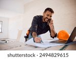 Happy construction worker communicating on cell phone while analyzing blueprints during home remodeling.