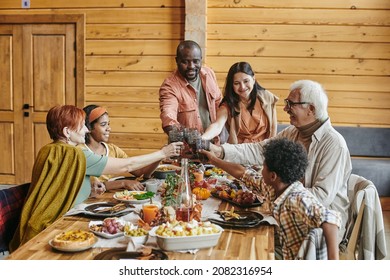 Happy contemporary interracial family clinking with drinks over served table while celebrating holiday in home environment