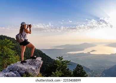A happy contented young girl with a backpack takes a souvenir photo of the Montenegrin coastal cities, the Adriatic Sea and the bright sunset rays from the top of a high mountain - Powered by Shutterstock