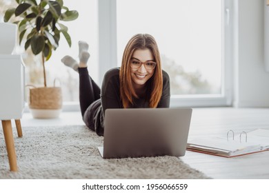 Happy Contented Young Woman Grinning To Herself As She Works On A Laptop Computer Lying In Her Socks On A Carpet In The Living Room In A Low Angle View