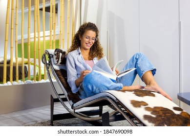 Happy Contented Young Woman Wearing Glasses Relaxing Barefoot On A Stylish Recliner Chair Reading A Book With A Smile