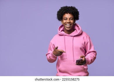 Happy cool African American teenage guy teen boy model wearing pink hoodie holding cell phone using mobile digital apps on cellphone pointing at smartphone isolated on light purple background. - Shutterstock ID 2255158275