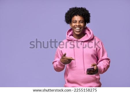 Happy cool African American teenage guy teen boy model wearing pink hoodie holding cell phone using mobile digital apps on cellphone pointing at smartphone isolated on light purple background.