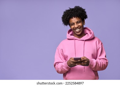 Happy cool curly African American teenage guy teen boy model wearing pink hoodie holding cell phone using mobile digital apps on cellphone texting on smartphone isolated on light purple background. - Shutterstock ID 2252584897