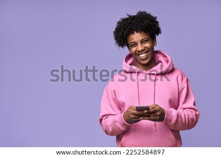 Happy cool curly African American teenage guy teen boy model wearing pink hoodie holding cell phone using mobile digital apps on cellphone texting on smartphone isolated on light purple background.
