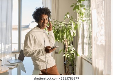 Happy cool gen z hipster African American guy wearing headphones holding phone at home, listening music or podcast, feeling relaxed standing in living room at home, portrait. - Powered by Shutterstock