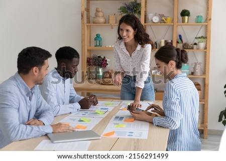 Happy corporate coach training diverse millennial team of interns, teaching marketing. Young business leader woman, mentor, boss talking to employees on meeting over sales reports