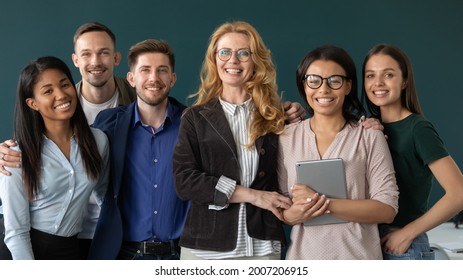 Happy corporate mature mentor, teacher, business coach and multiethnic group of interns, students looking at camera, smiling, hugging together, celebrating training course accomplishment. Head shot