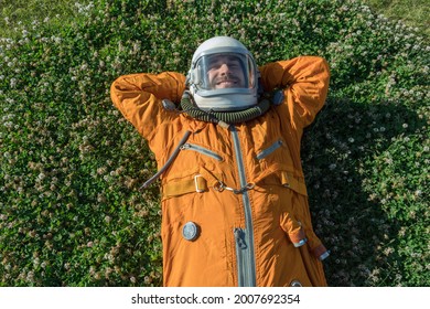 Happy Cosmonaut Wearing Orange Spacesuit Having A Rest While Lying On Green Grass