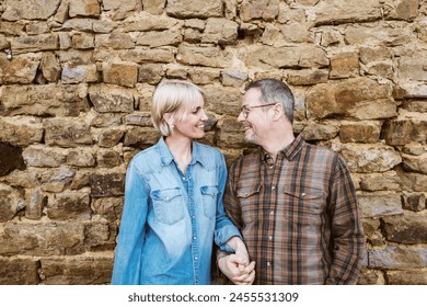 A happy couple, a 50-year-old man and a 40-year-old woman, stand outside in front of a stone wall, smiling and gazing into each other's eyes - Powered by Shutterstock