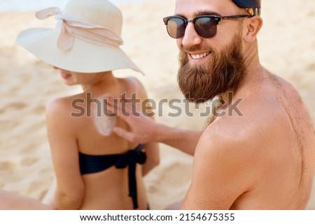 Happy Couple Applying Protective Sunscreen On Tanned Shoulder At Beach. Putting Sun Lotion On Female Skin. Skin And Body Care.
