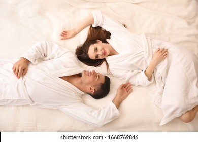 Happy couple in bathrobes lying on bed, top view