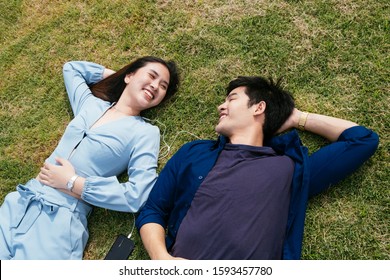 Happy Couple In Blue Clothes Lie Down On A Grass Field And Listening The Music Together.