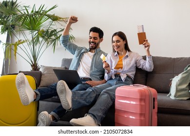 Happy couple booking hotel online using laptop and credit card, sitting with packed suitcases, passports and tickets, ready for vacation trip