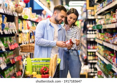 Happy Couple Buying Food In Supermarket, Choosing Products Standing With Trolley Cart Along Aisles And Full Shelves Purchasing Groceries Essentials Together. Smiling Spouses Holding Jar Of Sause - Powered by Shutterstock