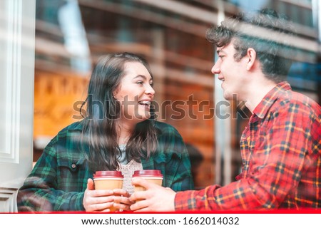 Happy couple in a cafe in London enjoying a coffee together - Man and woman holding a cup of tea or coffee and looking each other smiling and laughing - Photo taken through the glass window