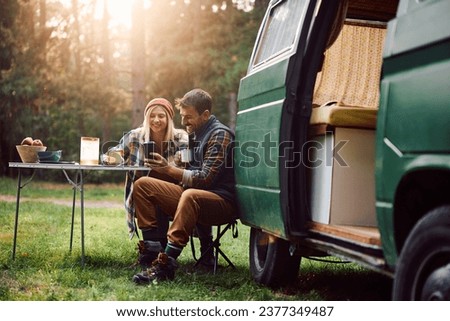 Happy couple of campers using cell phone while having a picnic at trailer park.