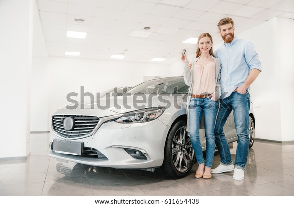 Happy couple with car key standing at car in dealership\
salon  