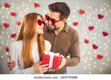 Happy couple celebrating Valentine's Day at home and enjoying spending time together and giving each other gifts. Couple in heart-shaped glasses posing on the background of a festively decorated wall.