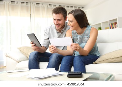 Happy Couple Checking Bank Account On Line In A Tablet Sitting On A Couch In The Living Room At Home