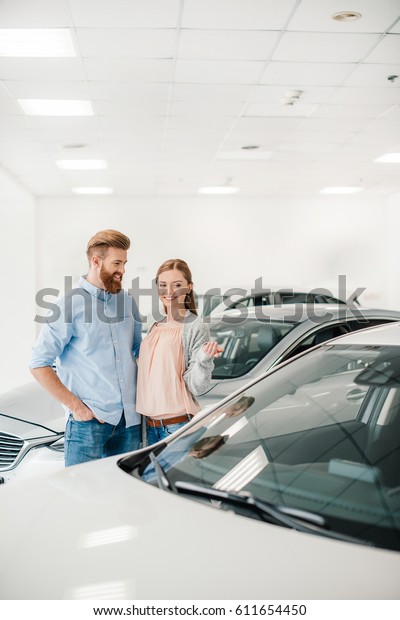 Happy couple choosing car in dealership salon, woman\
pointing on car