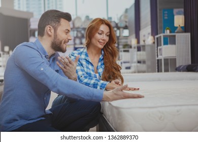 Happy Couple Choosing New Comfortable Mattress At Furniture Store. Beautiful Woman And Her Handsome Husband Examining Beds For Sale At Homeware Supermarket, Copy Space