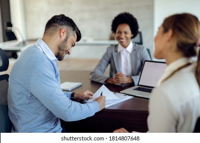 Happy Couple Closing A Deal With Their Bank Manager On A Meeting In The Office. Focus Is On Man Signing The Contract. 