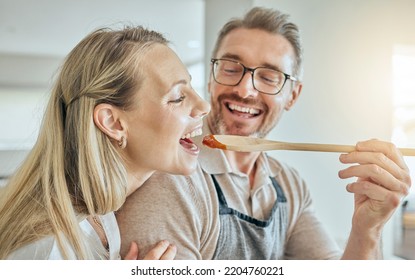 Happy Couple Cooking, Tasting Spoon Of Food And Meal, Lunch And Dinner In Kitchen At Home. Smile Mature Man Giving Hungry Woman A Taste Of Homemade Tomato Sauce Meal In Mouth To Enjoy Eating Together